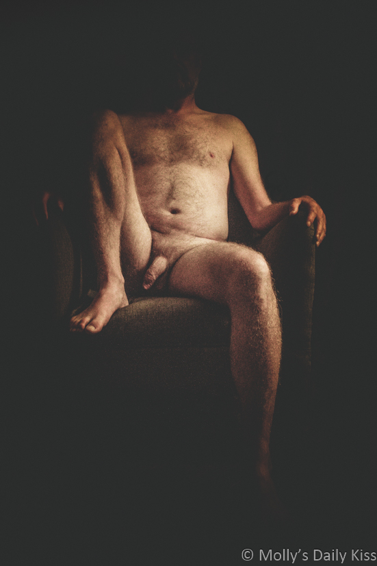 Nude of man sitting in chair with one leg up and his face hidden in the shadows