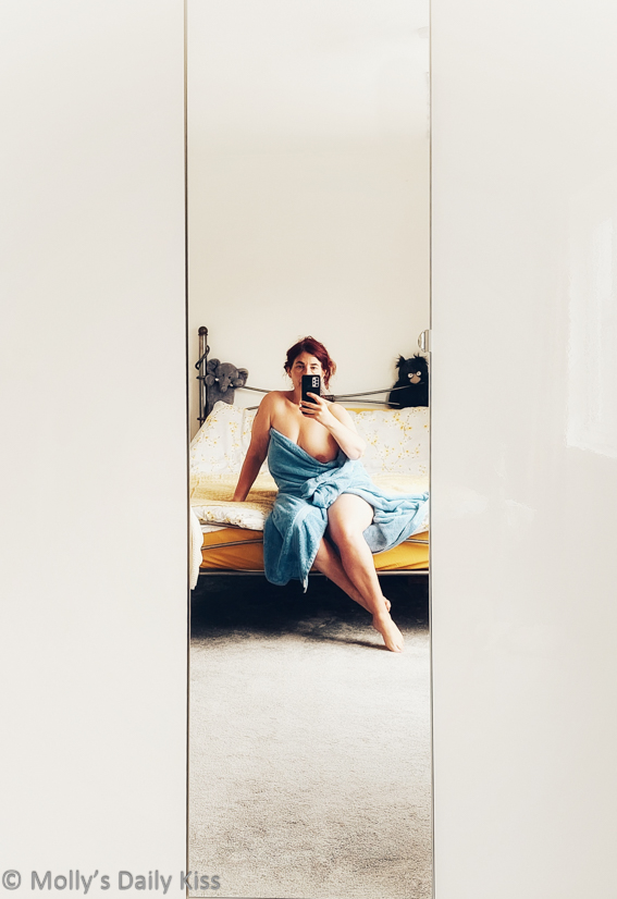 Molly sitting on the end of her bed in blue towel taking selfie in the full length mirror