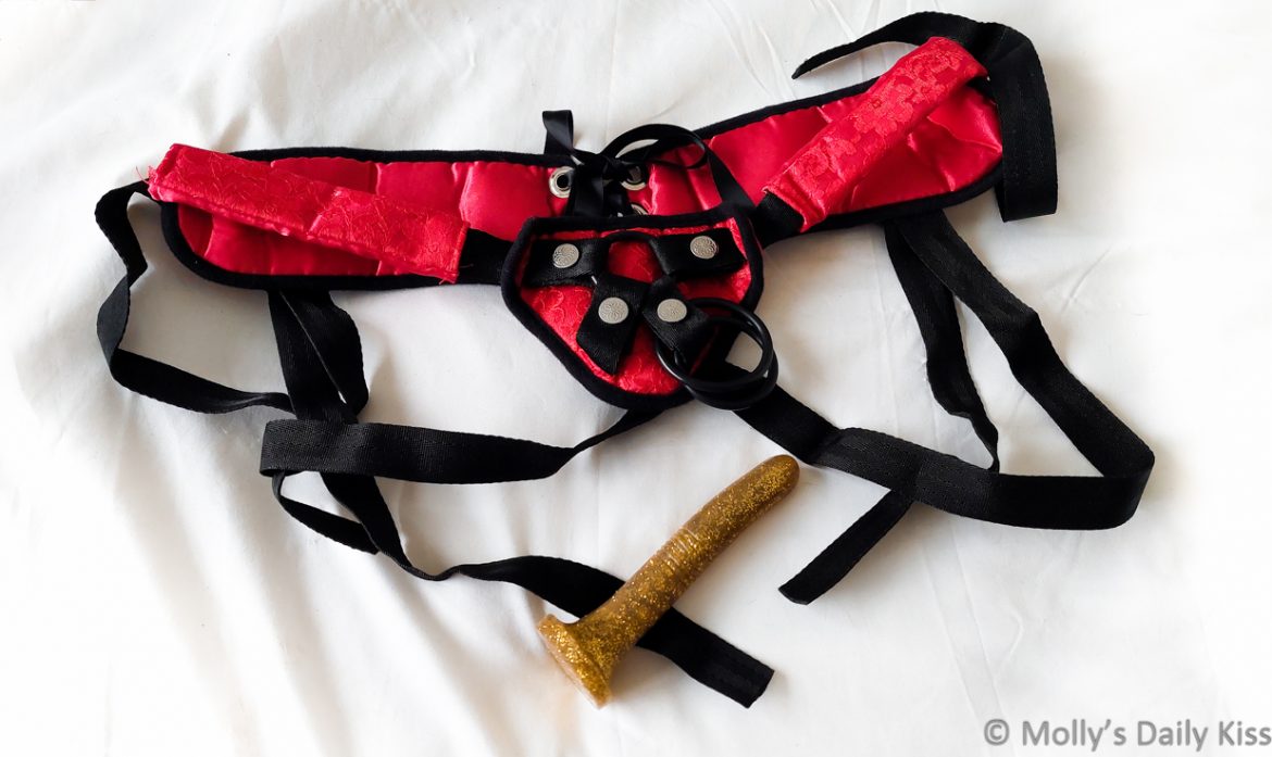 Red and black pegging strap-on harness and gold glitter dildo laying on the end of the bed