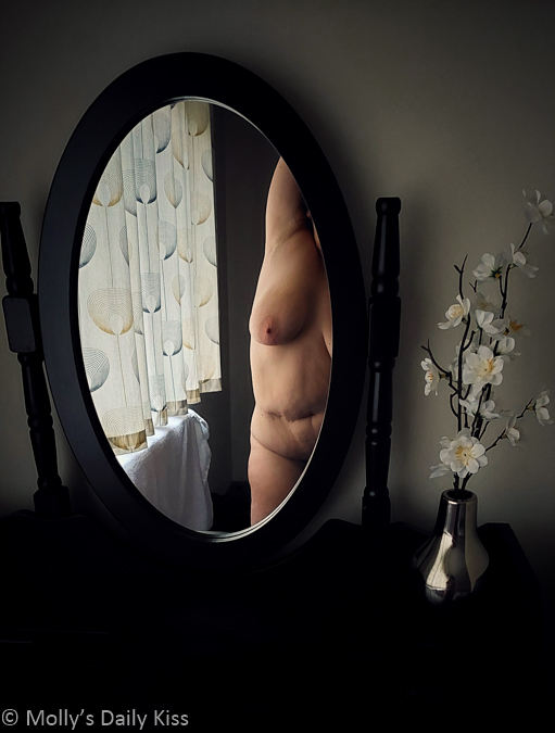 Oval dressing table mirror reflecting half of Molly's naked body showing the scar on stomach and one breasts, By the mirror is a little vase of white flowers for post called no reason to hide