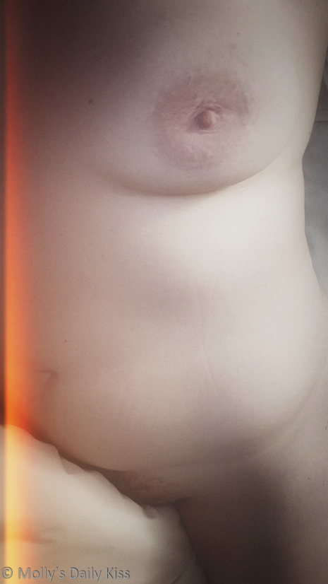 Looking down on mollys naked body. We can see one breast and the soft swell of her tummy and a bit of her pubic mound and top of her thigh. There is a cream coloured duvet covering most of her pubic mound and an orange light leak along the left of the image