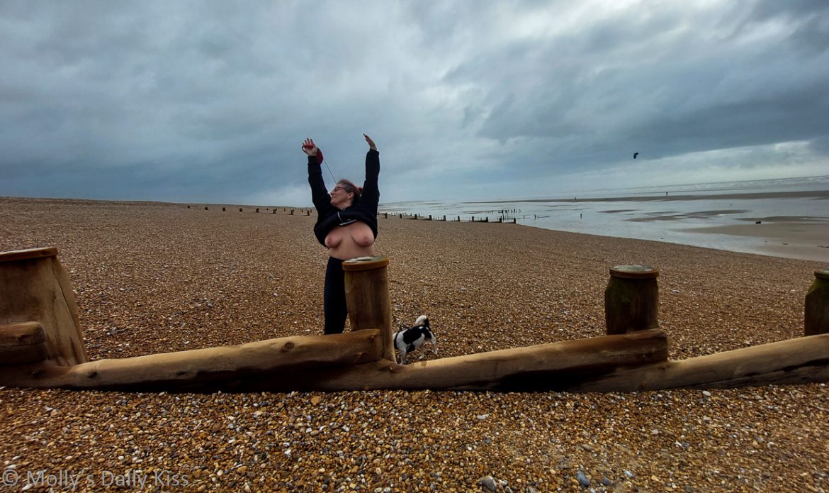 Groynes Molly standing on a peddle beach by the wooden groynes with her hands above her head and her top lifted up showing her boobs