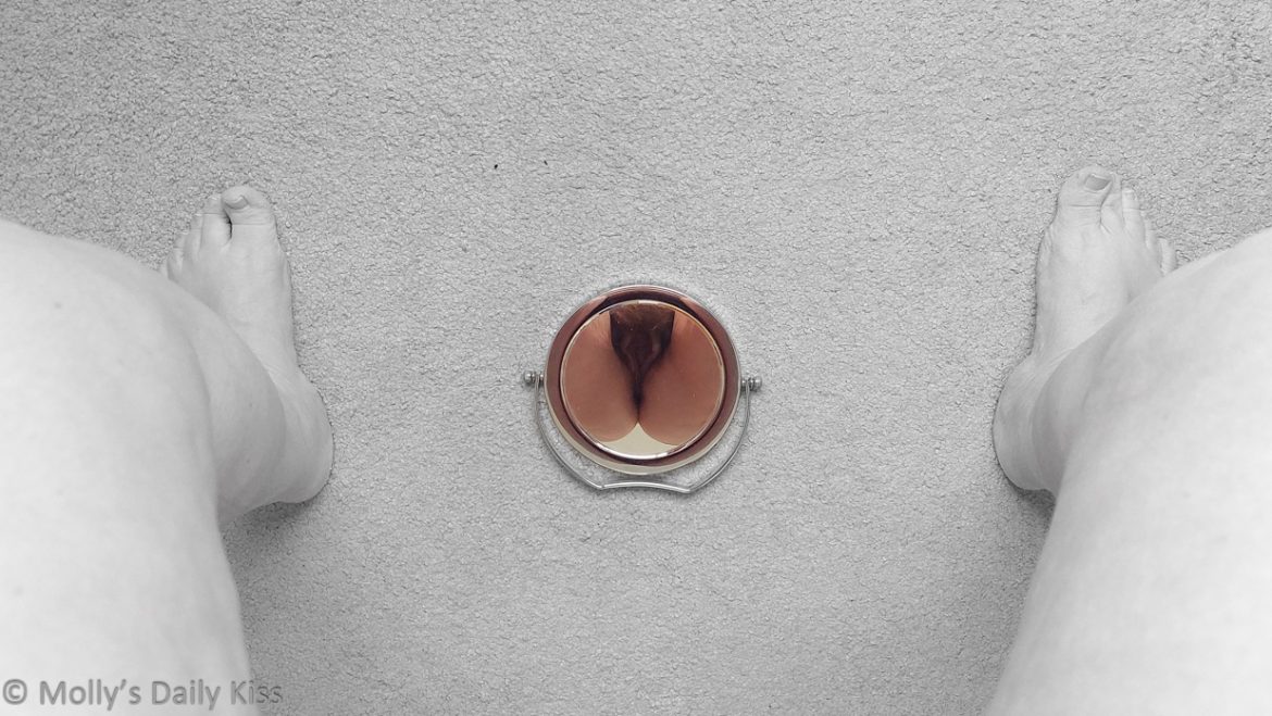 Looking down through molly's legs to a small round mirror on the floor. We can see her bare legs and feet and the carpet which are all in black and white. Her vulva is reflected in the small mirror and that part of the image is in colour. We can see her pubic hair and the vulva lips splaying out in a heart/oval shape