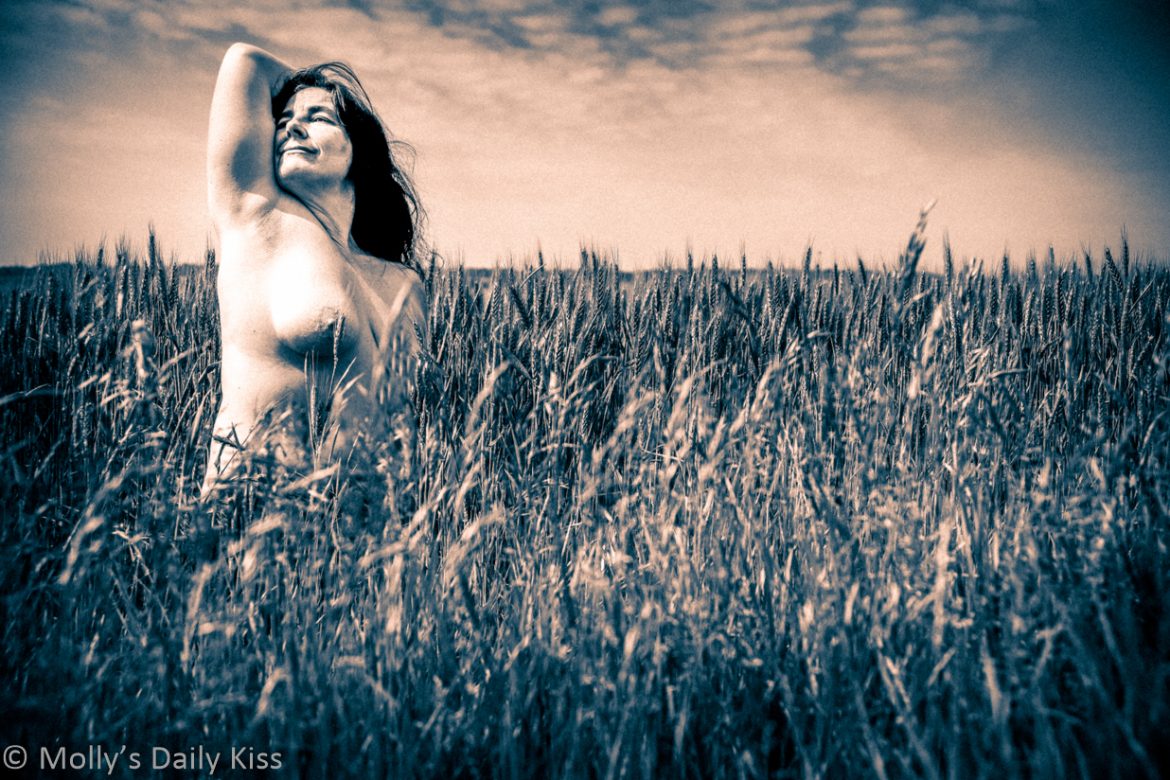 Molly topless in field of wheat crops. The image is edited to look like an old two tone movie with pale pinkish clouds and grey tones