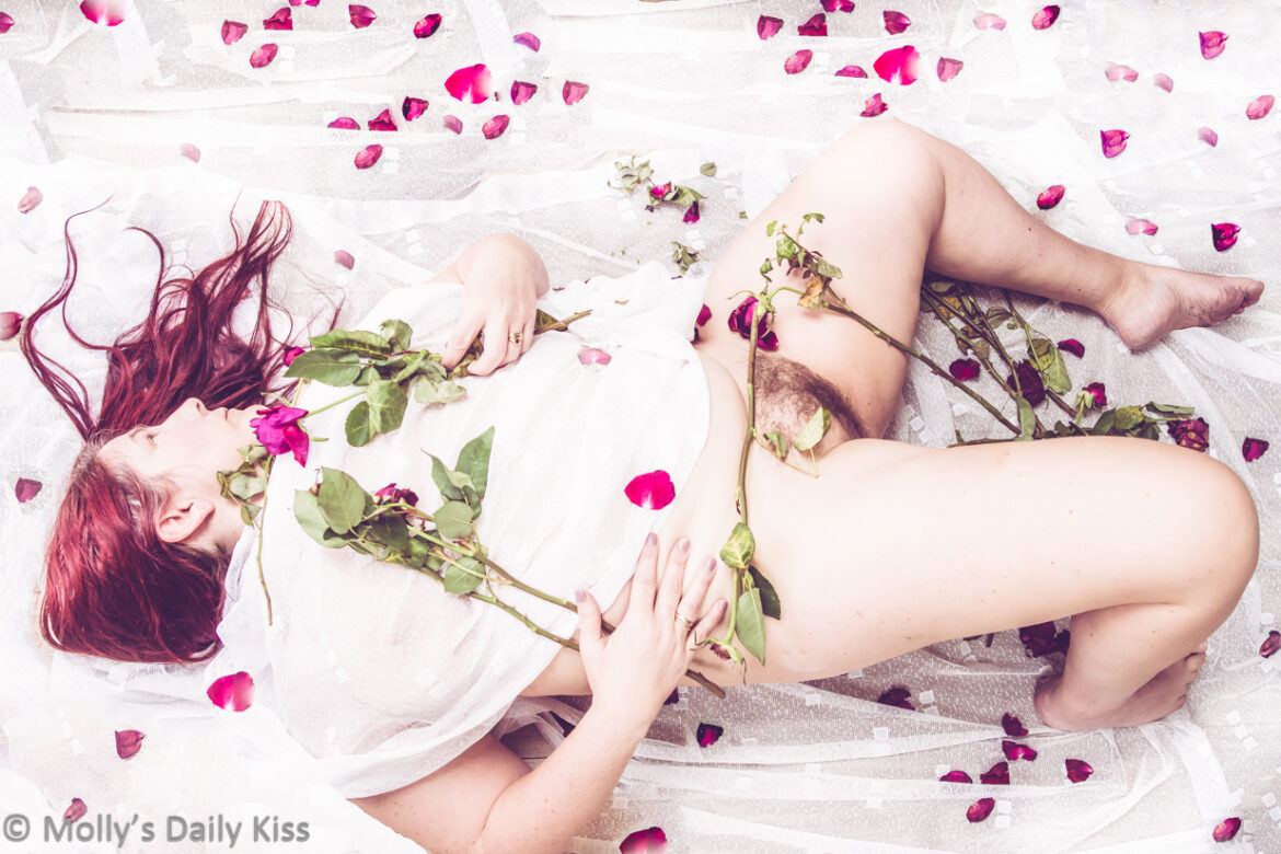 Looking down on molly laying on white material. She is holding wilted roses and there are rose stems on her leg and petals all round her. She has white material over her chest and belly but her legs are splayed open and we can see her pubic hair. Her red hair is splayed out round her head. The whites of the image are over exposed and her skin is a soft blush pink colour