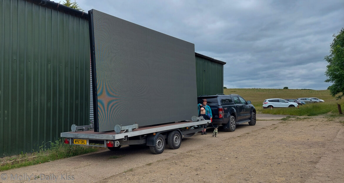 Molly flashing her breast next to large outdoor cinema screen that is on the back of a trailer