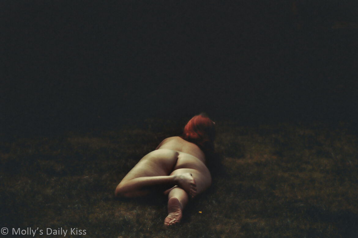 Molly laying naked on the grass with her back the camera. The image is dark but her skin is a bright pale tones and her red ahir pops. The image has a filter giving it a painting effect for post called Ariadne