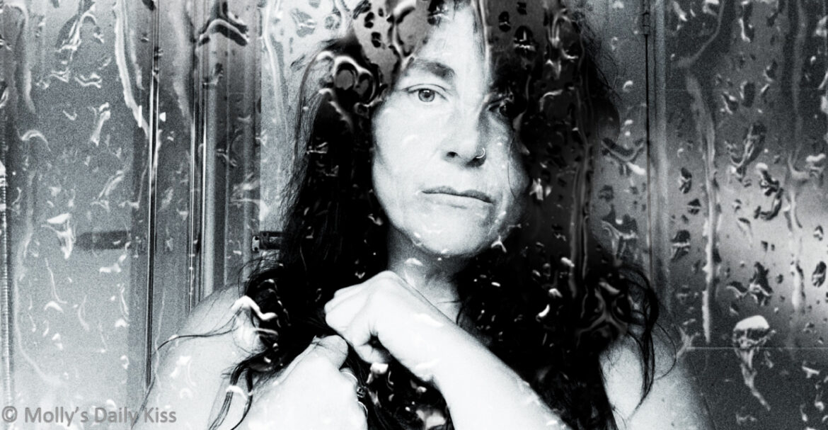 Cropped black and white of molly standing behind shower glass which is all wet. She is naked but clutching a towel to her chest. We can see one of her breasts and she looks serious/sad for post called maybe